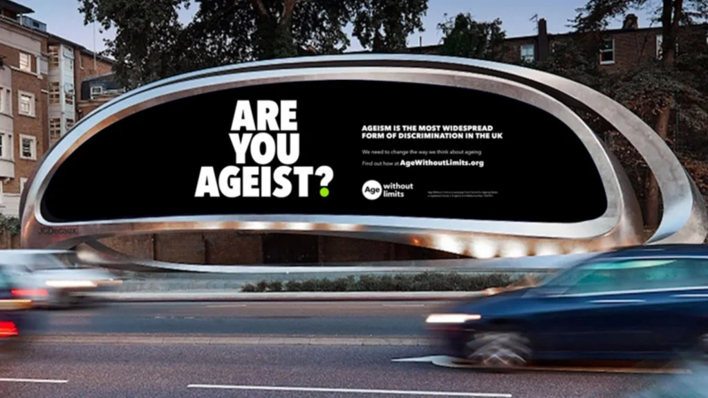 A roadside billboard saying Are You Ageist with cars driving past in front of it