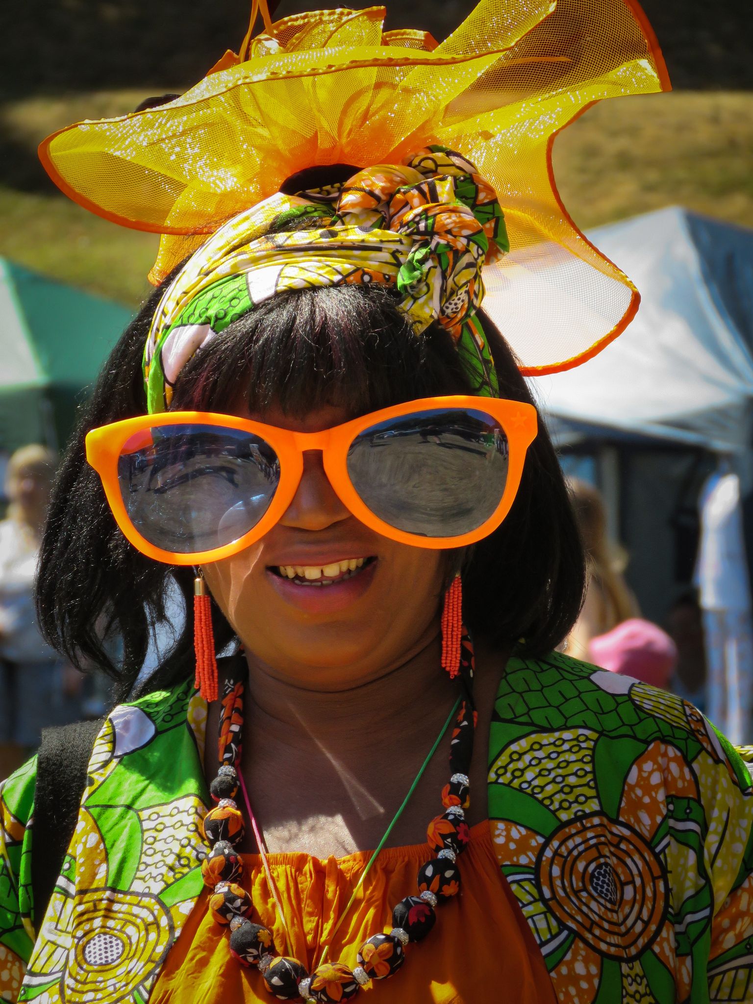 A woman smiles at the camera, wearing a large yellow hat and oversized orange sunglasses. The sun is shining on her.