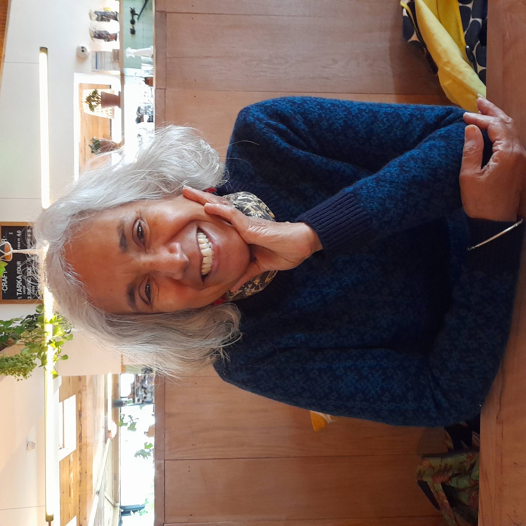 A woman with a silver grey bob sits at a table, resting on her arms and smiling.