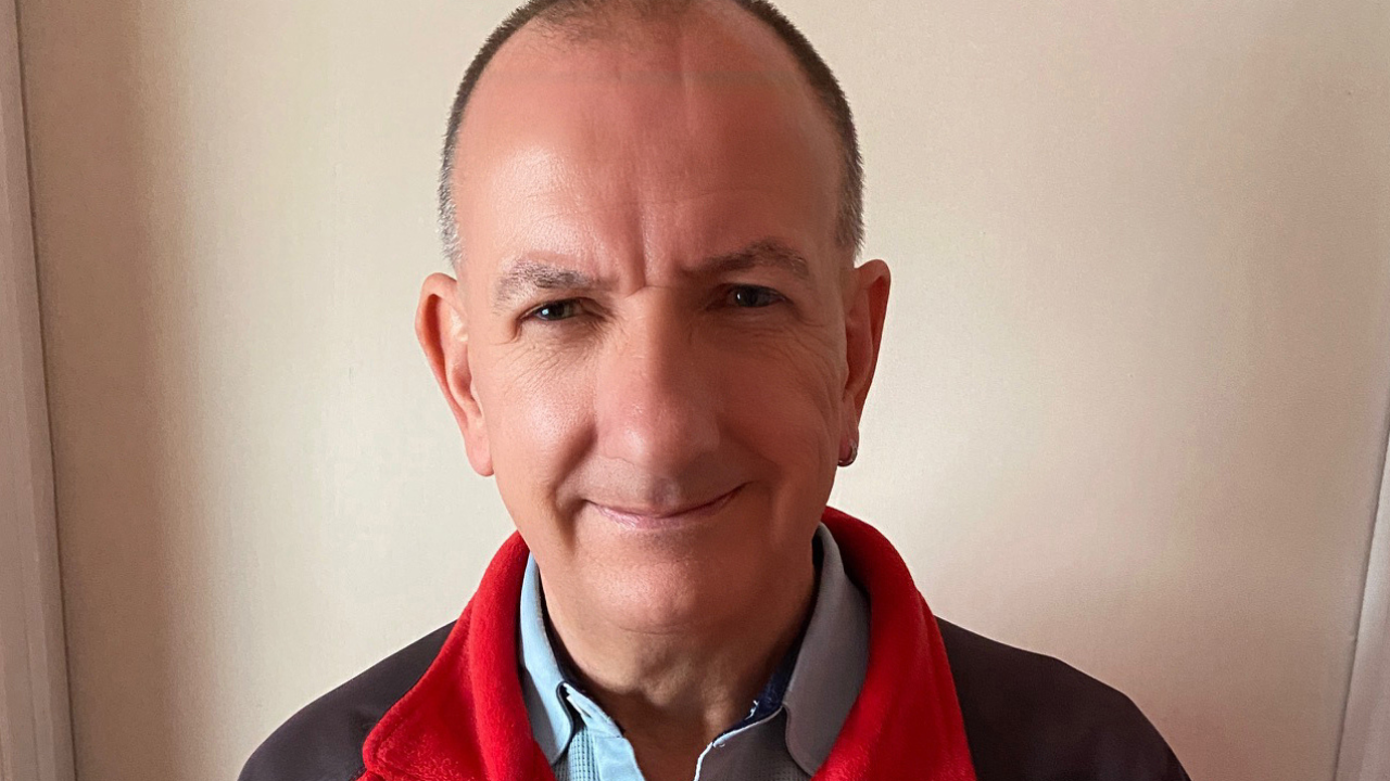 A man wearing a shirt underneath a fleece jacket smiles at the camera with a plain wall behind him.