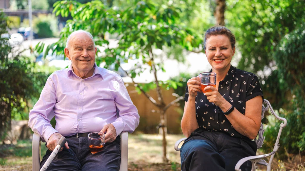 Ann and her father sitting outside holding cups of tea with sunny trees behind them.