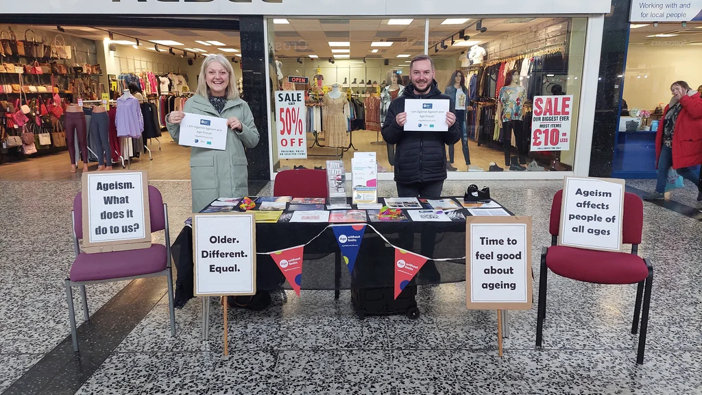 A man and woman stand behind a table displaying posters about ageism and Age Without Limits. Shops are in the background.