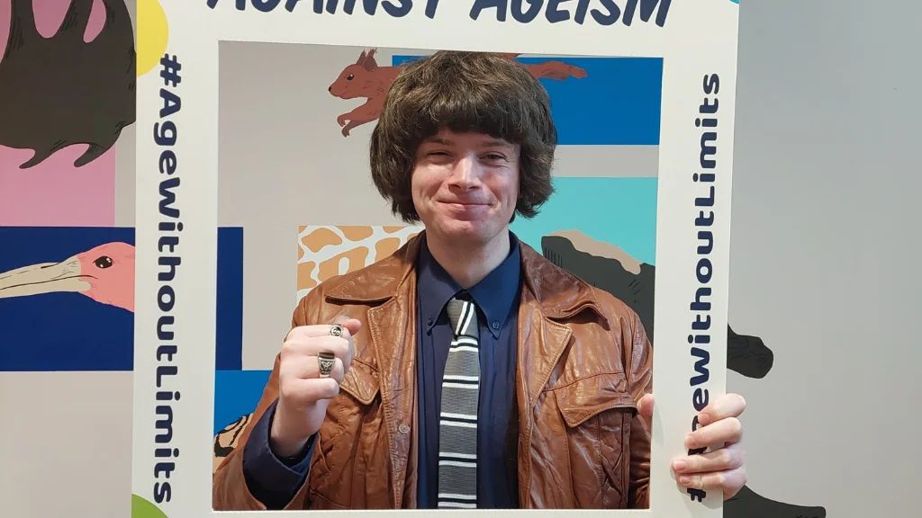 A young man holds a selfie frame which says Against Ageism, Age Proud.