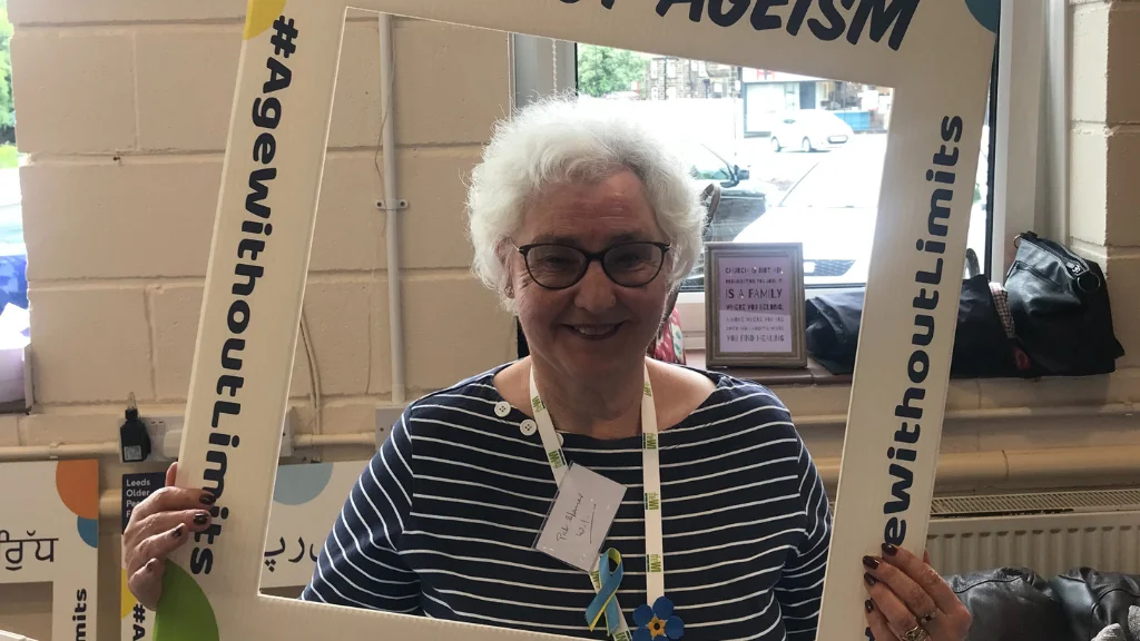 An older woman holds a selfie frame saying Against Ageism, Age Proud