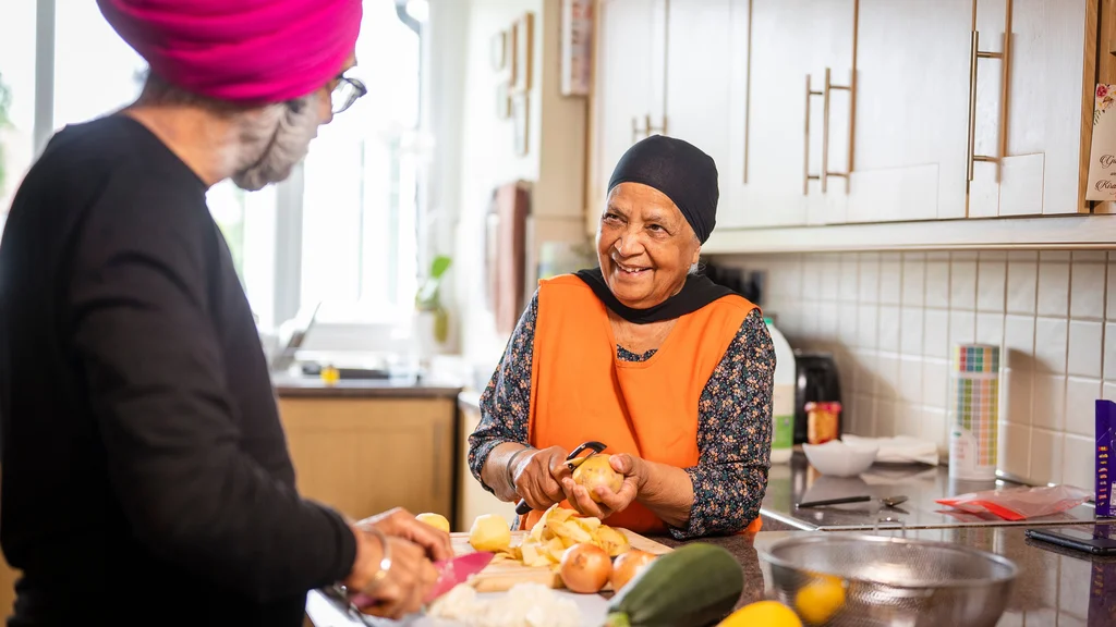 An older woman smiles at an older man while both chop vegetables in a kitchen.