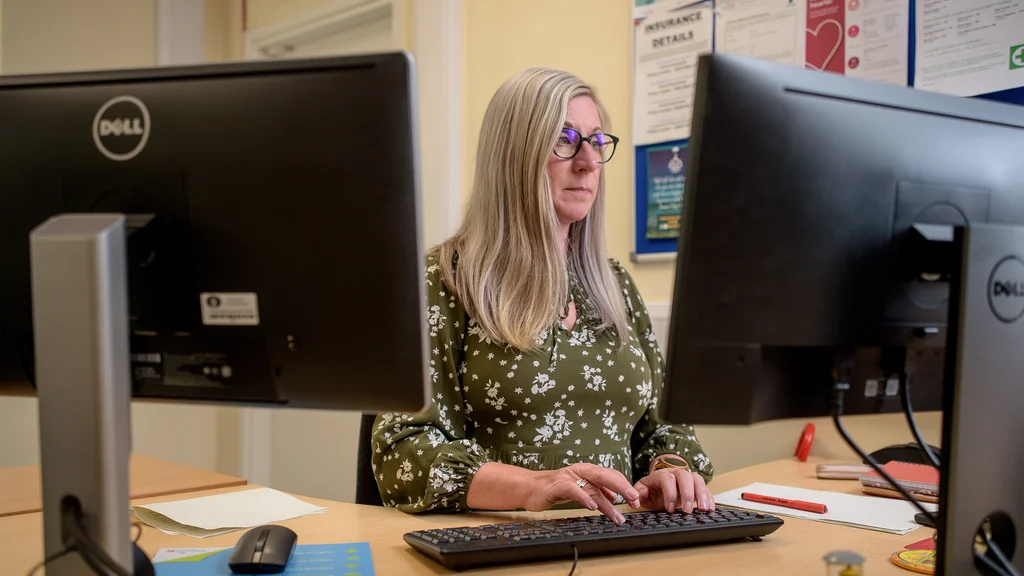 A woman sits at a desk behind two computer screens. She is typing on a keyboard.