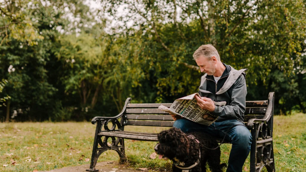 A man with grey hair sits on a bench with a dog and reads a newspaper.