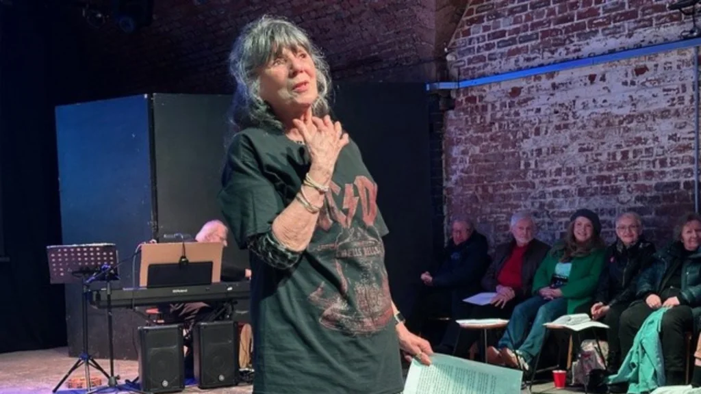 A woman holding a script and acting with audience members behind her.