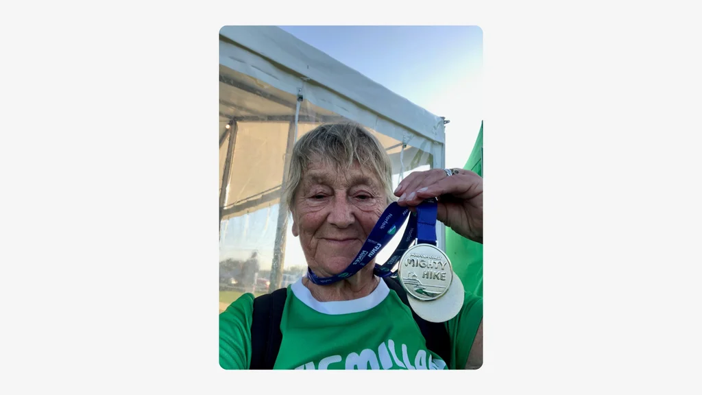 An older woman wears a Macmillan shirt and holds up a medal, smiling at the camera. Credit: Jeanne Smith.