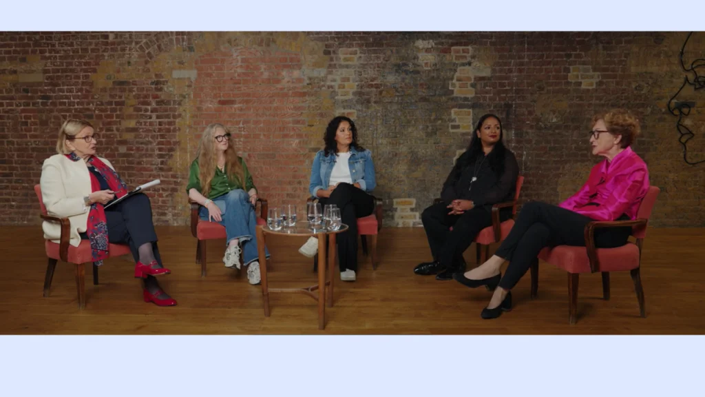 Five women talk while sitting on chairs in a row.