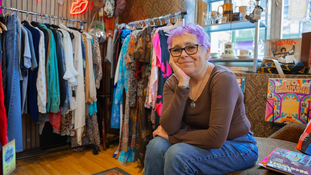 A woman with short purple hair sits on the edge of a couch and leans on her knees, smiling towards the camera. The background is full of colourful prints, objects and clothes.