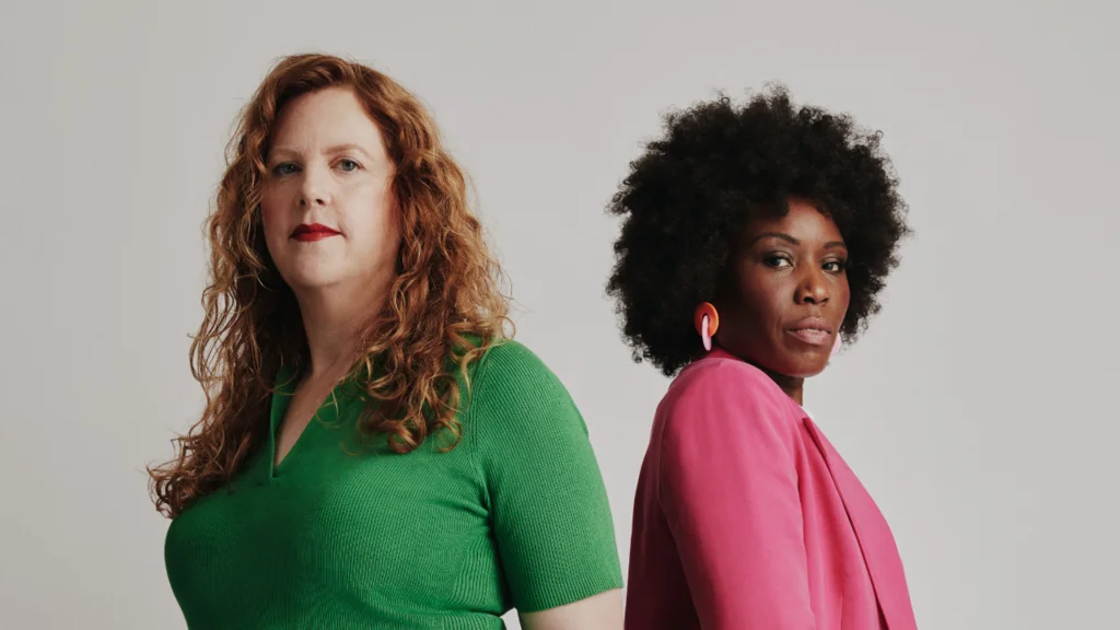 Two women, one dressed in bright green and one in bright pink, stand and look defiantly at the camera.