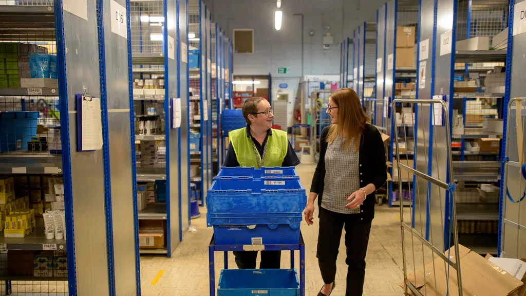 Two women speak while one pushes a trolly in a warehouse