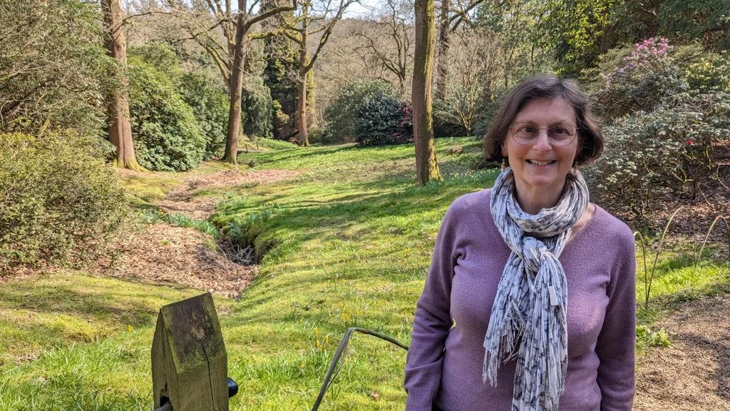 A woman wearing a lilac jumper and scarf stands outside surrounded by grass and trees