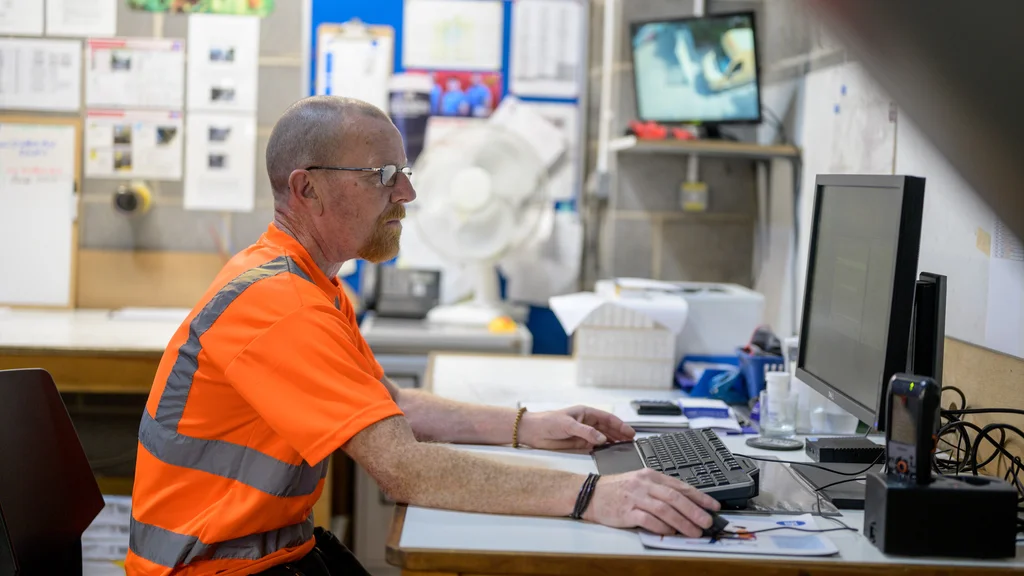 A man in a high vis t shirt works at a laptop at a desk