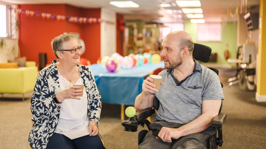 Two people drinking from disposable cups, with a table covered in balloons behind them