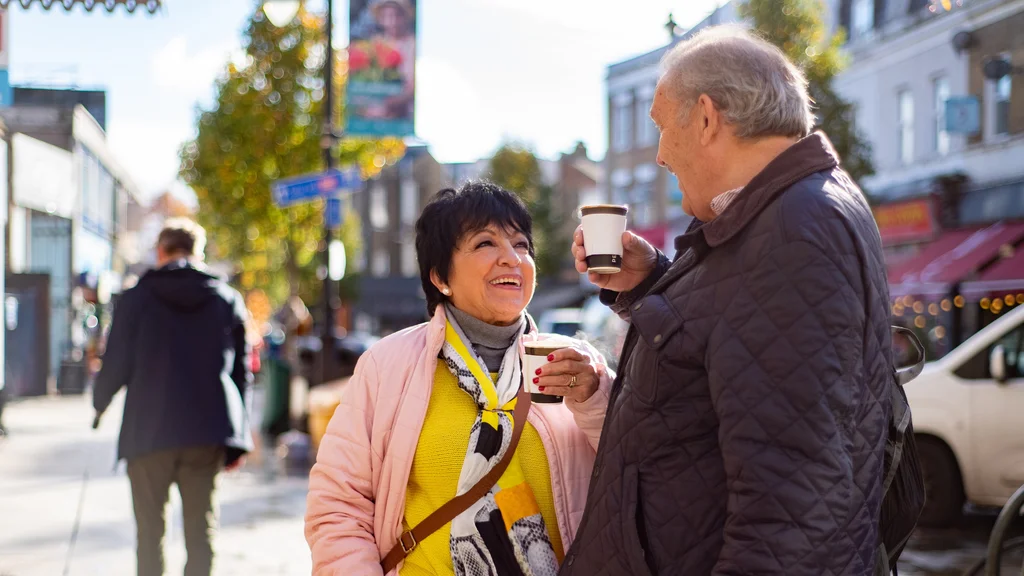 An older man and woman hold takeaway coffee cups and stand in a street